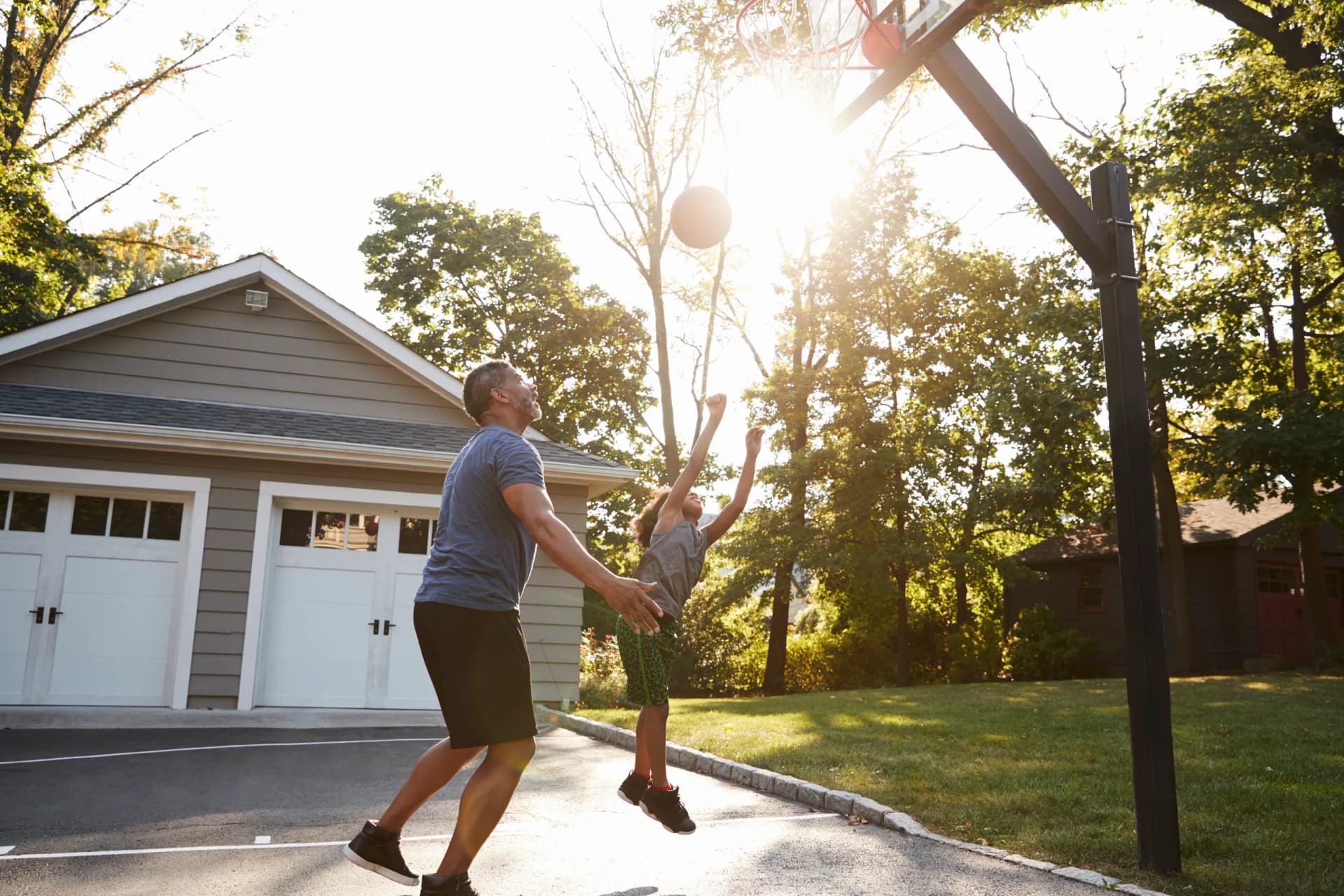 An adult man and a child playing basketball together on a sunny driveway outside a house.