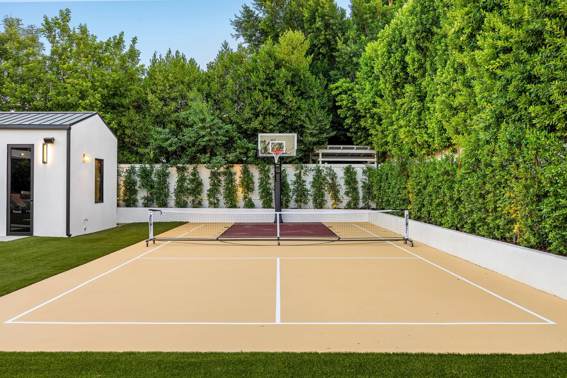 An outdoor multi-sport court with a pickleball/tennis net and a basketball hoop, next to a modern building and artificial grass, surrounded by lush hedges.